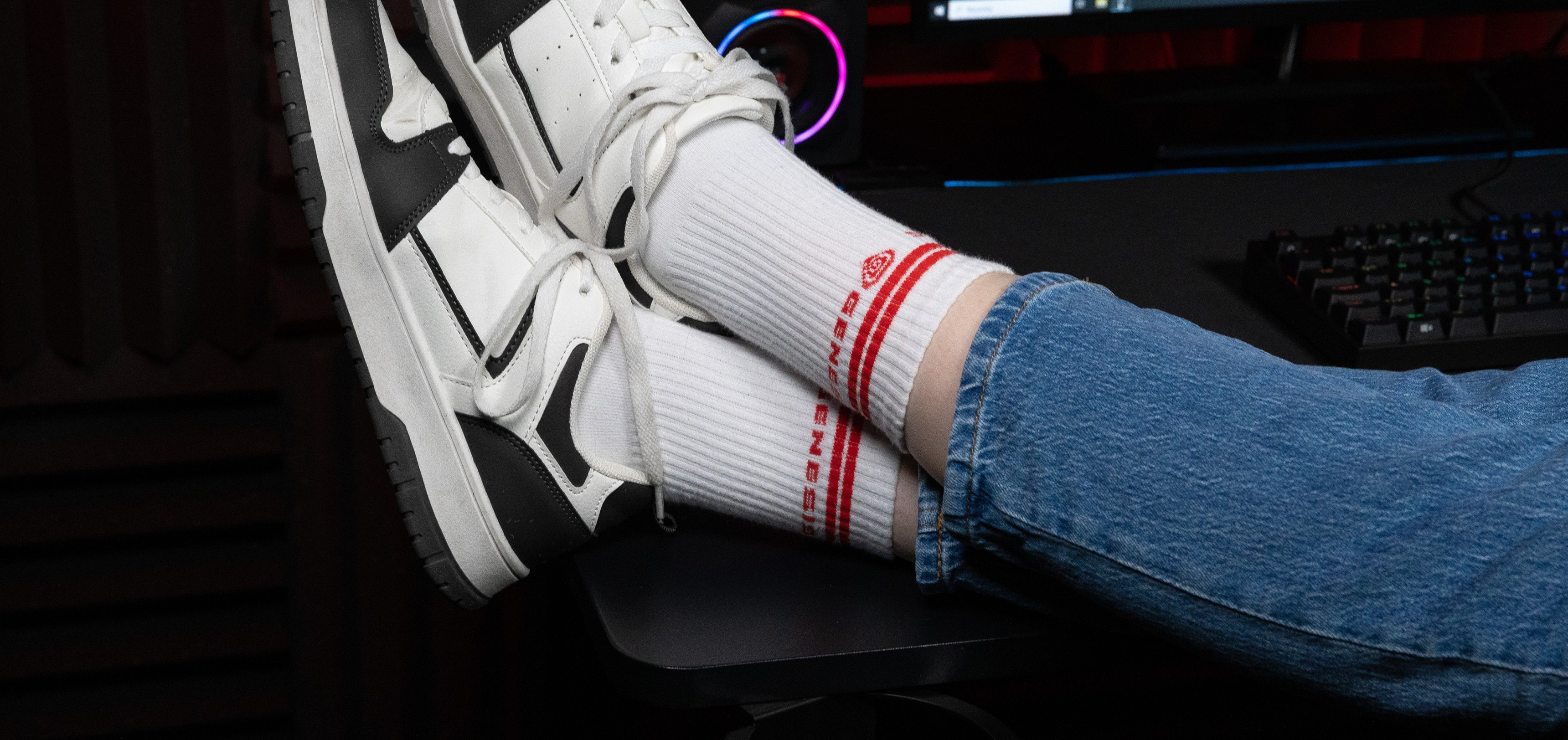 Chaussettes de sport blanches avec marquage rouge, portées avec des baskets blanches et noires, reposant sur un bureau avec une configuration informatique.